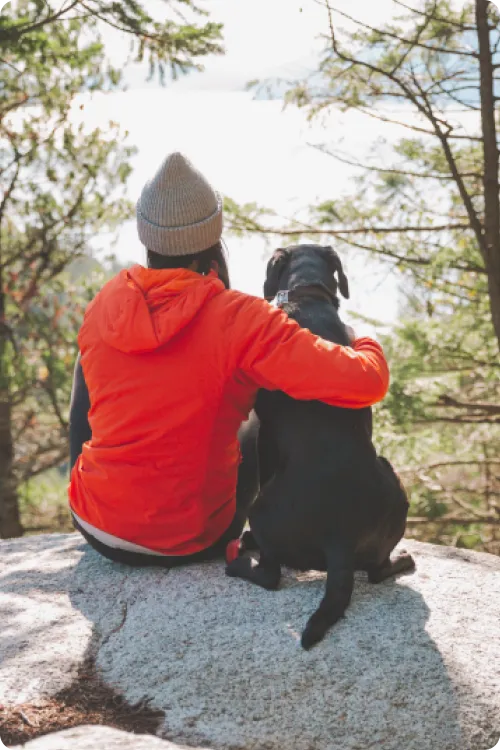 backview of lady in beanie with arm over black dog sitting on cliff looking at lake