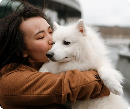 Une femme propriétaire d’animal de compagnie qui apprend l’assurance trupanion coûte embrasser leur chien blanc à l’extérieur.